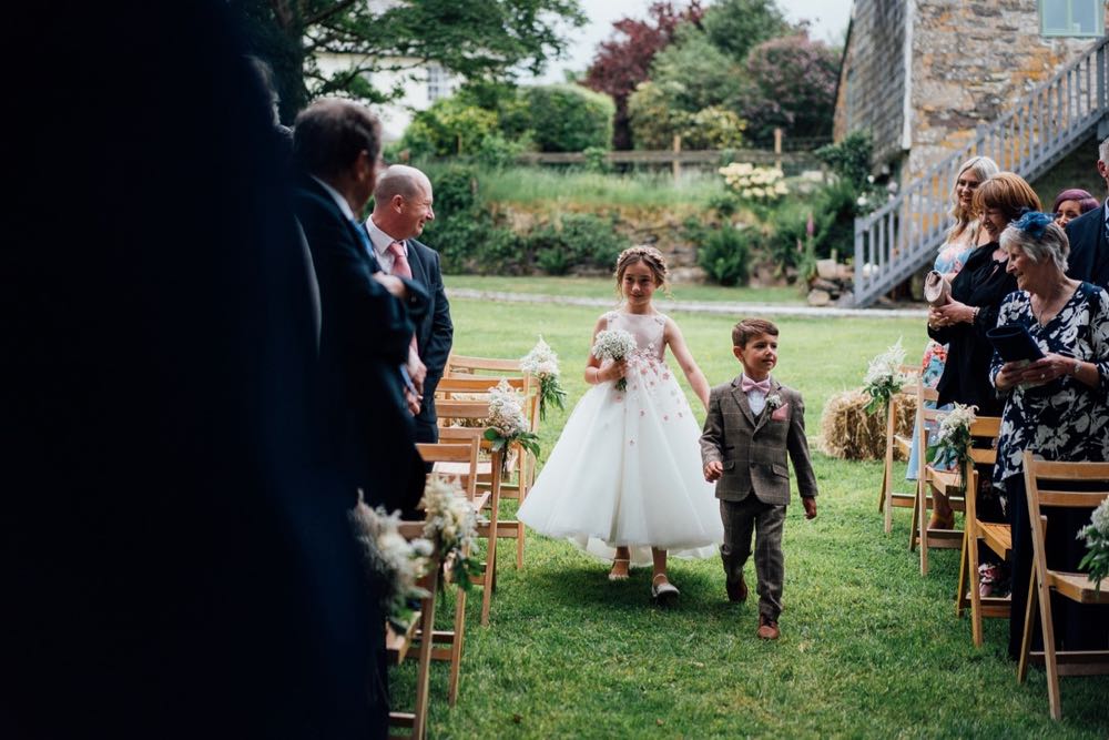 A flower girl and page boy walking down the \"aisle\" between 2 rows of chairs on the lawn