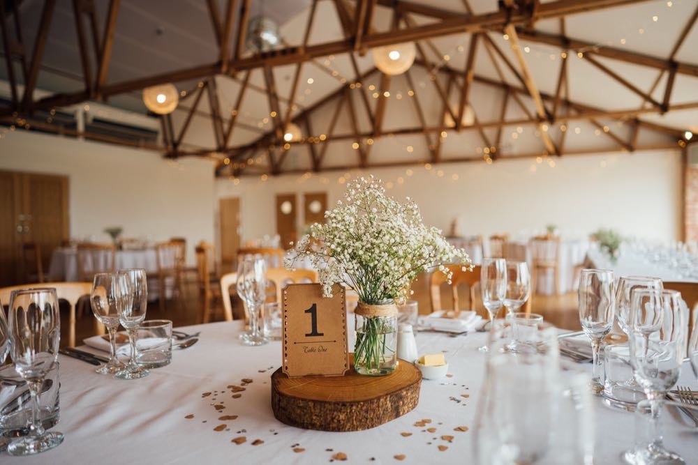 A shot of the inside of the Red Brick Barn set up for the wedding breakfast, There are round tables with white cloths. The centrepieces are log slices with jars of gyp