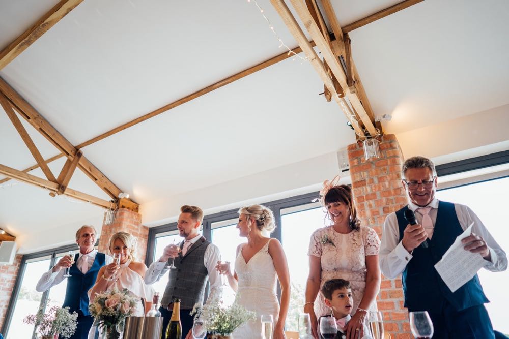 A picture taken in the Red Brick Barn. The picture is of the guests on the top table. Teh top table is set up in front of the windows. The guests are standing for a toast