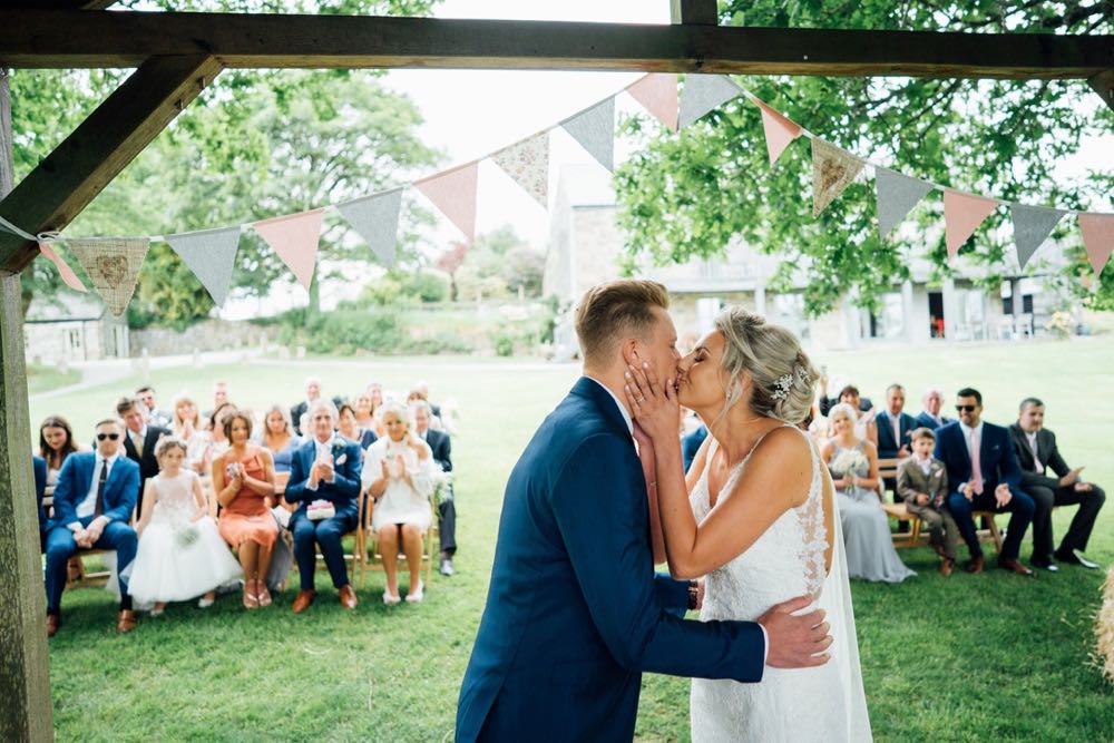 \"You may kiss the bride\" moment. The shot is taken from the back of the arbour with the couple in the foreground. It is taken from the waist up. The groom is  on the left and the bride is on the right. The groom wears  dark grey suit and the bride wears a white sleeveless dress. She has blond hair work up.
