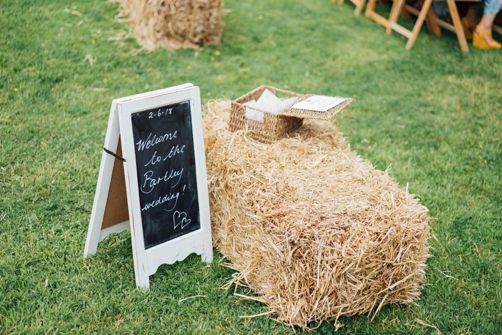 A decor detail shot showing a straw bale on the lawn and a hand written a-fram chalk board. Th e sign reads \" Welcome to the Bartley wedding\"