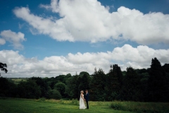 A shot of the couple with the Cornish landscape in the background. The couple are tiny compares to the background. There is a line of fir trees ion the horizon and the sky is blue with some clouds