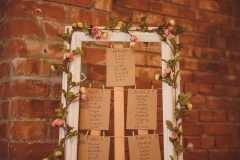 This is an image of a table plan against a red brick wall. The table plan is framed with white picture frame entwined with pink roses. Each table is repersented by a buff card that has names written on it
