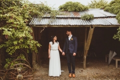 A bride and groom stand in front of an open fronted shed. The bride is on the left. They are holding hands and looking at each other. The bride is wearing a long, white sleeveless dress and has her hair loose. The groom is wearing a dark, suit, white shirt and brown brogues.