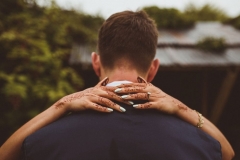 This omage is of the back of the grooms head and shoulders. He is wearing a blue suit jacket and has short mid-brown hair. The bride's hands are around his neck and they are decorated with henna.