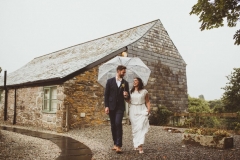 A bride and groom are walking in front of a granite stoen barn. The bride is on the right. They are holding hands and looking at each other and are under an umbrella.. The bride is wearing a long, white sleeveless dress and has her hair loose. The groom is wearing a dark, suit, white shirt and brown brogues. They are walking towards the camera.