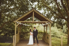 A bride and groom stand under the Oak Arbour. The bride is on the left. They are holding hands and looking at each other. The bride is wearing a long, white sleeveless dress and has her hair loose. The groom is wearing a dark, suit, white shirt and brown brogues.
