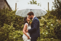 A bride and groom kiss under an umbrella in front of lots of greenery. The bride is on the left.  The bride is wearing a long, white sleeveless dress and has her hair loose. The groom is wearing a dark, suit, white shirt and brown brogues.