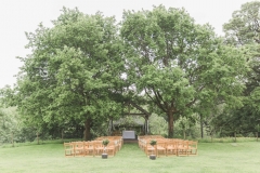 The Oak Arbour in May with each tree in full leaf. A full length shot showing the arbour set between 2 large oak tress. Wooden chairs are set out in the grass in frot of the arbour and the trees are in full leaf.