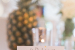 A close up shot showing the detail of the table decor. There is a large  pineapple in the background sitting on a log slice. The table linen is white and there is a table place name set in a wooden holder. The sign says \"BRIDE\".