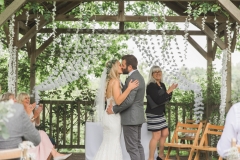 This is a picture of the bride and groom under the arbour  after the registrar has said \"you may kiss the bride\". The couple are facing each other and kissing. The bride is on the left. She is  wearing a white, fitted, sleeveless dress with shoestring straps. She has blond hair that she is wearing loose. She is wearing  a flower garland and a veil. The groom is wearing a light grey suit. He has dark hair and a beard