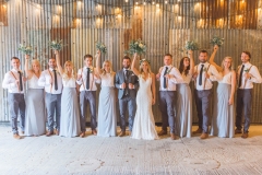 This is a picture of the bridal party in the really Rustic Barn. They ar standing in front of the corrugated iron clad wall. The group is made up of the bride and groom, 5 bridesmaids and 6 groomsmen. The bridesmaids are wearing long grey skirts and white blouses. The groomsmen are wagon brown loafers, grey trousers, blue ties, white shirts and red braces. Each bridesmaid and the bride is raising her bouquet into the air. The photo shows them in full length and there is festoon lighting above them.