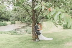 This is a picture of the bride and groom standing under a cherry tree by the lake.  About two thirds of the tree is shown and the couple look quite small next to it.  The couple are facing each other and kissing. The bride is on the right. She is  wearing a white, fitted, sleeveless dress with shoestring straps. She has blond hair that she is wearing loose. She is wearing  a flower garland and a veil. The groom is wearing a light grey suit. He has dark hair and a beard