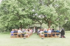 The Oak Arbour in May with each tree in full leaf. A full length shot showing the arbour set between 2 large oak tress. Wooden chairs are set out in the grass in frot of the arbour and the trees are in full leaf. Guests are seated in the chairs and the bride and groom are under the arbour.