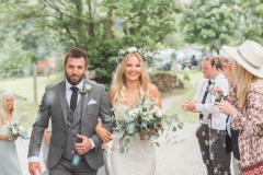 This is a picture of the bride and groom walking towards the Green Room after the cermeonyarbour. The bride is on the right. She is  wearing a white, fitted, sleeveless dress with shoestring straps. She has blond hair that she is wearing loose. She is wearing  a flower garland and a veil. The groom is wearing a light grey suit. You can see guests standing beside them. They are linking arms and walking towards the camera. the shot is from the knees up. There are trees and stone barns in the background.
