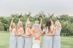 This is a picture of the bride and bridesmaids standing on the lawn in front of the Red Brick barn. They are standing in front of the camera with their back to it and you can see a view of a green landscape in the background. The bride is in the middle with 2 bridesmaids on either side. The bridesmaids are wearing long grey skirts and white blouses.  Each bridesmaid and the bride is raising her bouquet into the air. The photo shows them from the knees up,