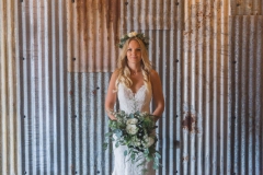 This is a picture of the bride in the really Rustic Barn. She is standing in front of the corrugated iron clad wall.  The photo shows her  from the knees up. She is  wearing a white, fitted, sleeveless dress with shoestring straps. She has blond hair that she is wearing loose. She is wearing  a flower garland and a veil and is holding a bouquet.