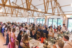 This is an image of the Red Brick Barn set up with long trestle tables. The centrepieces are pineapples on log slices. You can see the bar on the left hand side and the rustic beams in the ceiling. Guests are seated at the tables on wooden Cheltenham chairs