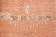 This is a close up of some decor details. It shows a red brick wall. There is a ledge in the centre of the shot. On the ledge there are letters saying \"H & S\" and some jam  jars with greenery. Above the shelf there is a dream catcher in white. Below the shelf there is some bunting made up of feathers.