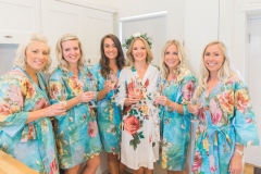 This is a picture of the bride and her 4 bridesmaids getting ready. They are standing in a row and wearing matching floral knee length gowns. The bridesmaids gowns are turquoise and the bride\'s is white. The bride is wearing a floral crown