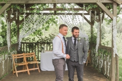 This is a picture of the bride and best man under the arbour waiting for the bride to arrive. The registrars table and 2 folding wooden chairs can be seen in the background.  The groom is on the righ. and is wearing a light grey suit. He has dark hair and a beard. The best man is dressed similarly but is not wearing a jacket or waistcoat. He has red braces. They are both looking towards where the bride will come from.