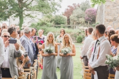 This is a picture of two of the bridesmaids walking down the \"aisle\" towards the arbour. They both have blond his that they are wearing loose. The hold bouquets in front of them. They are wearing grey skirts and white blouses. You can see guests standing beside their chairs on either side of the aisle. There are trees and stone barns in the background.