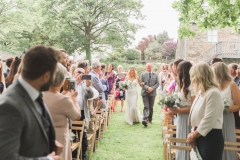 This is a picture of the bride and the person who is giving her away walking down the \"aisle\" towards the arbour. The bride is on the left. She is  wearing a white, fitted, sleeveless dress with shoestring straps. She has blond hair that she is wearing loose. She is wearing  a flower garland and a veil. The man is wearing a light grey suit. You can see guests standing beside their chairs on either side of the aisle. There are trees and stone barns in the background.