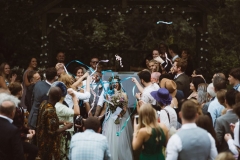 This is taken just after an outdoor ceremony under the Oak Arbour. It is summer and the trees ae in ful leaf. The bride is on the right and the groom on the left.  The image is taken from the knees up with the Oak Arbour framing the shot. The couple are walking away fromthe arbour towards the camera and their guests are facing them. The guests are throwing ribbon cenfetti at them and the couple are laughing.