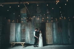 An imageof the couple taken in the Really Rustic Barn. The couple are standing and the bride is on the left and the groom on left. There are strings of festoon lighting above them