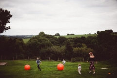 This shot shows some guests on the lawn with orange spacehoppers. There is a wood covered hill in the background.