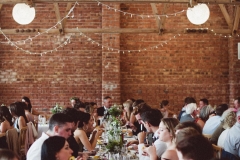 A group of guests are sitting at  trestle table in front of a Red Brick wall.