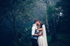The couple are embacing in the woods. The light is dark and they are in a pool of light. The bride is on the right and the grrom on the left.