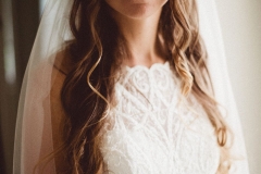 Thia is a close up, head and shoulders shot of the beautiful bride. She is wearing a a white sleeveless dress with lace details. She has long dark hair that she is wearing loose. She is smiling and looking at the camera.