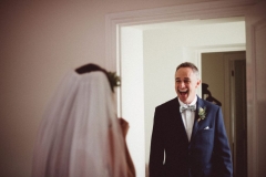 This picture shows the bride's father seeing her in her wedding dress for the first time. They are both shown from the waist up. You can see the back of the brde and you can see the faceof thefather of the bride. He is open-mouthed and very happy. The bride wears a veil. The fatherof the bride weara a navy suit, white shirt and bowtie