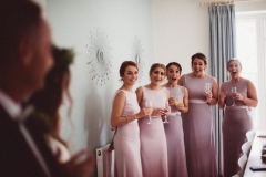 This image shows the 5 bridesmaids seeing the bride in her derss for the first time. The bride and her father are in the far left of the shot shown from the side. The image is focussed on the breidesmaids. The are facing the camera, wearing pink, sleeveless, full-length gowns. They are holding chanpagne  glassses. Their faces are full of joy.