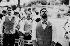 This image has the groom in the middle. He is waiting at the Oak Arbour with his back to the the guests. He is wearing a white shirt, a bowtie and a tweed waistcoat. This is a black and white shot. You can see 2 of his groomsmen behind him and they are similarly dressed. The groom looks really nervous and is looking upwards.