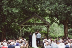 This is taken during an outdoor ceremony under the Oak Arbour. It is summer and the trees ae in ful leaf. The guests are seated in two colums of rows of chairs in front of the arbor with their abcks to the camera. The couple are under the arbour with thir back to the camera with the bride on the left and the groom on the right.  The image is taken from a distance with the Oak Arbour framing the shot.