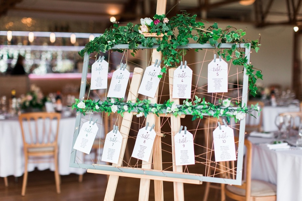 This picture is taken in the Red Brick Barn, It shows a detail shot of the couple's table plan, The table is made of a wooded picture frame that is draped in greenery. There are buff tags tied to the table plan and each is the name of a table with the guests names on it. In the background the Red Brick barn is set up with round tables with white linen and cheltenham chairs.