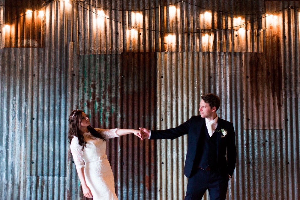 The bride and the groom are standing in the middle of the Really Rustic barn there is festoon lighting in the ceiling . The couple are shown only from the knees up. There is a corrugated iron wall behind them.  The bride and groom are standing in the middle of the shot and are standing side by side holding hands. The bride is on the left and is leaning away from he groom, She wears a fitted white gown with lace details and a delicate floral crown. The groom wears a dark suit with a white shirt and tie.