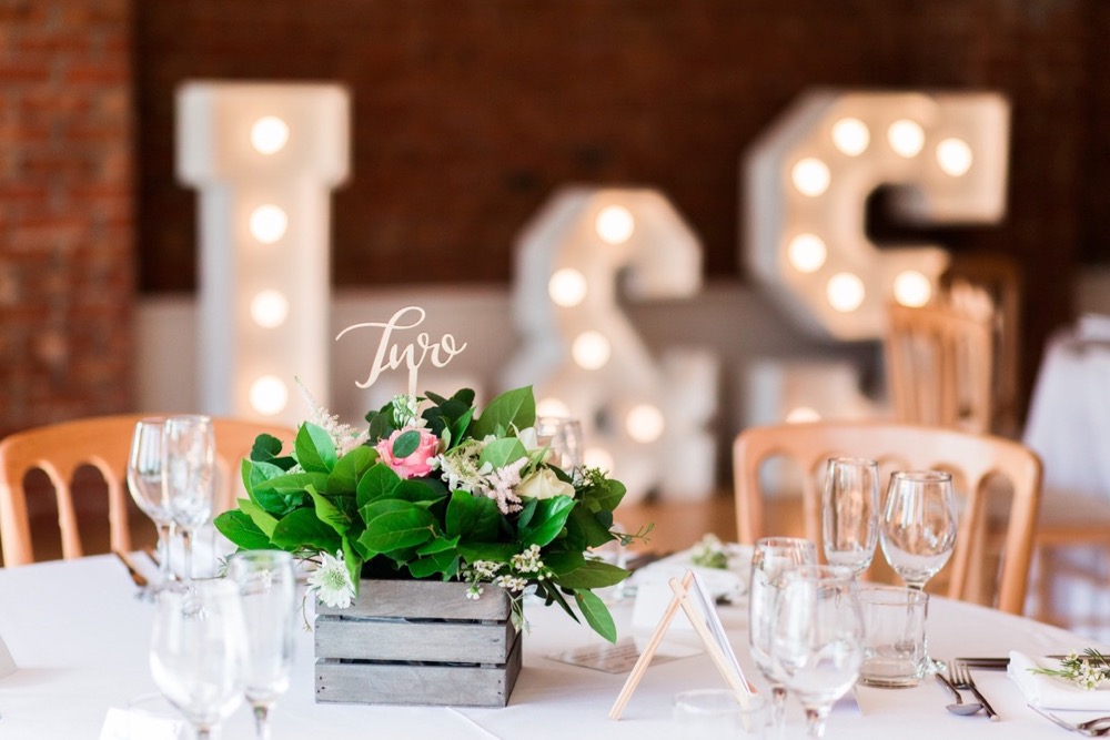 Table set up with round tables, white linen, cheltenham chairs and floral centre-pieces. Light up letters with L & S in the background.