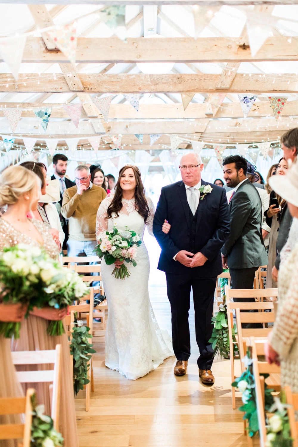 The bride and her father walk down the isle. The bride is on the left. She wears a fitted white gown with lace details and a veil. the pic is takes from the front of the wedding  barn and you can see bunting on the beams. The FOB wears a dark suit with a white shirt and a bow-tie. The bride holds a bouquet of white and pink flowers and it is hand-tied. There is a bridesmaid in the foreground to the left who holds a similar bouquet. The guests are standing bedside their chairs looking at the bride and FOG. Wooden folded chairs are set out in two columns of rows with an aisle down the middle.