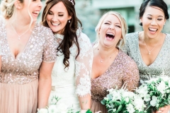 This is an image of the bride with 3 of her bridesmaids. It is a close up shot and they are laughing. There are 4 girlish the shot. The bride is second from the left. She wears a fitted white gown with lace details and 3/4 length lace sleeve.  She wears a delicate floral crown.  The bridesmaids are wearing  rose pink dresses with cap-sleeves and sequin embellishment.