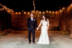 The bride and the groom are standing in the middle of the Really Rustic barn there is festoon lighting in the ceiling and much of the barn is in shadow. There are red brick walls and a rough concrete floor.  The bride and groom are standing in the middle of the shot and are standing side by side holding hands. The bride is on the right. She wears a fitted white gown with lace details and a delicate floral crown. The groom wears a dark suit with a white shirt and tie.
