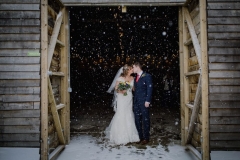 A full length shot of the bride and groom standing at the entrance to the Really Rustic Barn. They are standing side by side and facing forwards but turning towards each other to kiss. The doors of the barn are open and they tower above the couple. You can see snow falling against the dark interior of the barn. The bride is wearing a white cap sleeved dress . She has dark hair that she is wearing loose with a veil. . The groom is wearing a dark grey suit.