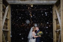 A waist up shot of the bride and groom standing at the entrance to the Really Rustic Barn. They are standing side by side and facing forwards but turning towards each other to kiss.The bide has her bouquet in her right hand. The doors of the barn are open and they tower above the couple. You can see snow falling against the dark interior of the barn. The bride is wearing a white cap sleeved dress . She has dark hair that she is wearing loose with a veil. . The groom is wearing a dark grey suit.