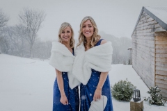 A picture of two bridesmaids standing outside in the snow. The shot is from the knees up. There is lots of snow in the background. The bridesmaids both have blonde hair that they are wearing loose. There are wearing blue dresses and white fur stoles