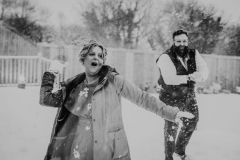 A back and white shot of two wedding guests having a snowball fight. The woman is wearing a cost and is covered in snow. The man is bearded and is in shirt sleeves.