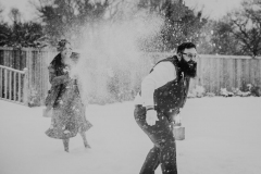 A back and white shot of two wedding guests having a snowball fight. The woman is wearing a cost and is covered in snow. The man is bearded and is in shirt sleeves.