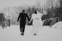 A full length shot of the bride and groom walking away from the camera outside in the snow. . They are walking side by side and facing forwards but turning towards each other. The bride is on the right. The bride is wearing a white cap sleeved dress . She has dark hair that she is wearing loose with a veil. . The groom is wearing a dark grey suit.