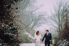 A shot from the knees up of the bride and groom walking away from the camera outside in the snow. . They are walking side by side and facing forwards but turning towards each other. The bride is on the left. The bare winter trees are towering above them. The bride is wearing a white cap sleeved dress . She has dark hair that she is wearing loose with a veil. . The groom is wearing a dark grey suit.